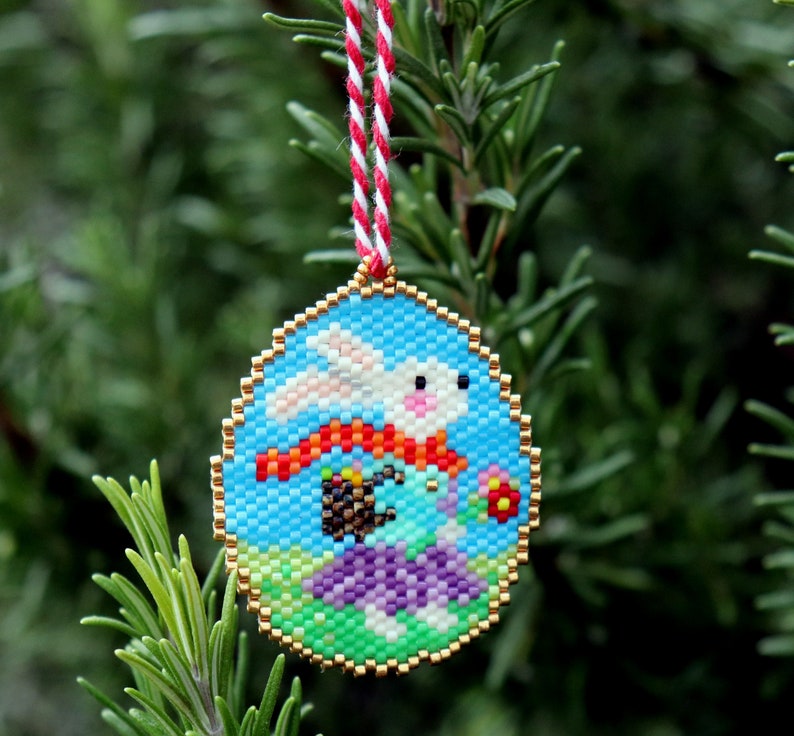 May include: A beaded bauble featuring a white bunny with a red scarf, holding a basket of colourful eggs. The bunny is on a green and purple background with a blue sky.
