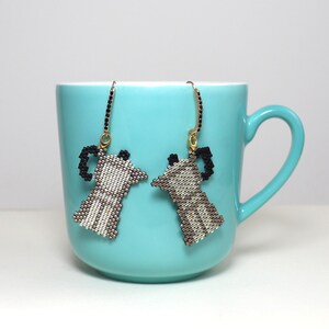 May include: A pair of beaded earrings shaped like coffee makers, in shades of white, brown, and black, hang in front of a turquoise mug. The earrings have gold-colored hooks and are a unique accessory for coffee lovers.