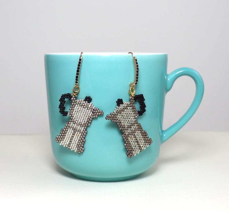 May include: A pair of beaded earrings shaped like coffee makers, in shades of white, brown, and black, hang in front of a turquoise mug. The earrings have gold-colored hooks and are a unique accessory for coffee lovers.