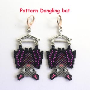 Könnte beinhalten: Ein Paar Perlen-Fledermaus-Ohrringe. Die Ohrringe zeigen ein schwarzes und lila Fledermaus-Design, das an einem silberfarbenen Kleiderbügel hängt. Der Körper der Fledermaus ist grau mit rosa Akzenten. Der Text "Pattern Dangling bat" befindet sich darüber.