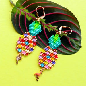 Puede incluir: Un par de pendientes de pi&ntilde;a de cuentas coloridas con detalles dorados. Cada pendiente tiene un peque&ntilde;o dije de palmera y un dije de flamenco rosa.
