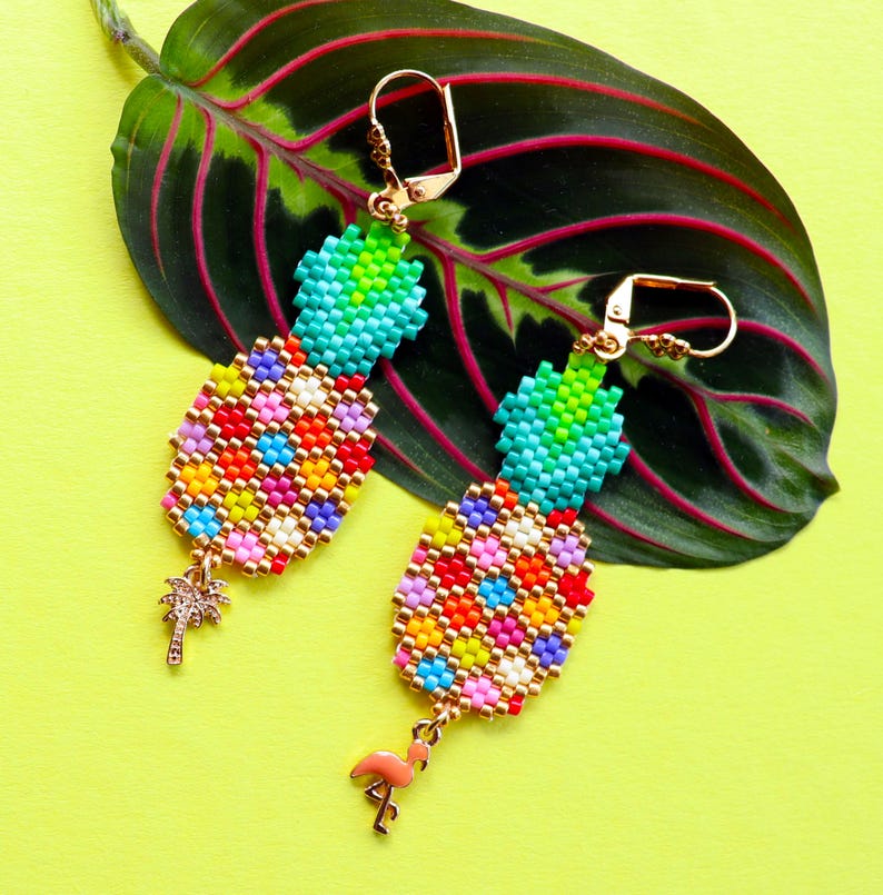 Puede incluir: Un par de pendientes de pi&ntilde;a de cuentas coloridas con detalles dorados. Cada pendiente tiene un peque&ntilde;o dije de palmera y un dije de flamenco rosa.