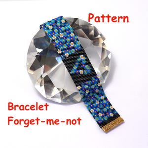 Könnte beinhalten: Ein schwarzes und blaues Perlenarmband mit einem Vergissmeinnicht-Blütenmuster. Das Armband liegt auf einer durchsichtigen Kristalloberfläche. Der Text "Pattern" befindet sich über dem Armband und der Text "Bracelet Forget-me-not" unter dem Armband.