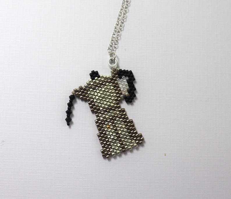 May include: A beaded pendant necklace featuring a coffee maker design. The pendant is made of small, metallic beads in shades of silver, brown, and black. The necklace has a silver chain and a lobster clasp.