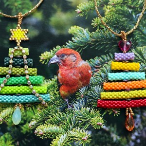 May include: Two beaded Christmas tree pendants with gold chains hang against a backdrop of evergreen branches. One tree is green with a yellow star, the other is rainbow-coloured. A red bird perches on the branches between the pendants.