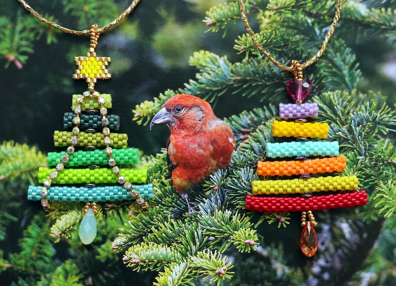 May include: Two beaded Christmas tree pendants with gold chains hang against a backdrop of evergreen branches. One tree is green with a yellow star, the other is rainbow-coloured. A red bird perches on the branches between the pendants.