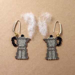 May include: A pair of handmade earrings featuring a coffee pot design. The earrings are made with small beads in shades of black, white, and gray, with gold-colored ear wires. Fluffy white material is arranged above the earrings to resemble steam.