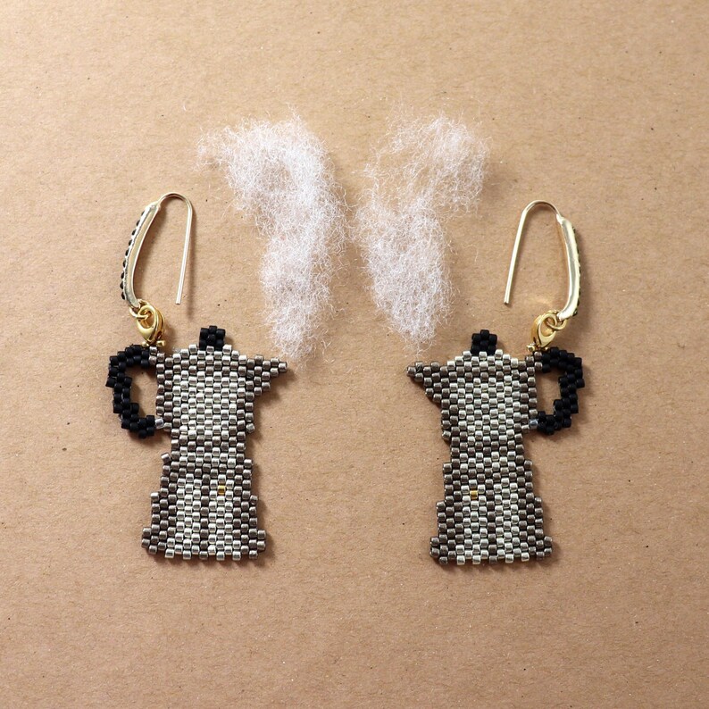 May include: A pair of handmade earrings featuring a coffee pot design. The earrings are made with small beads in shades of black, white, and gray, with gold-colored ear wires. Fluffy white material is arranged above the earrings to resemble steam.