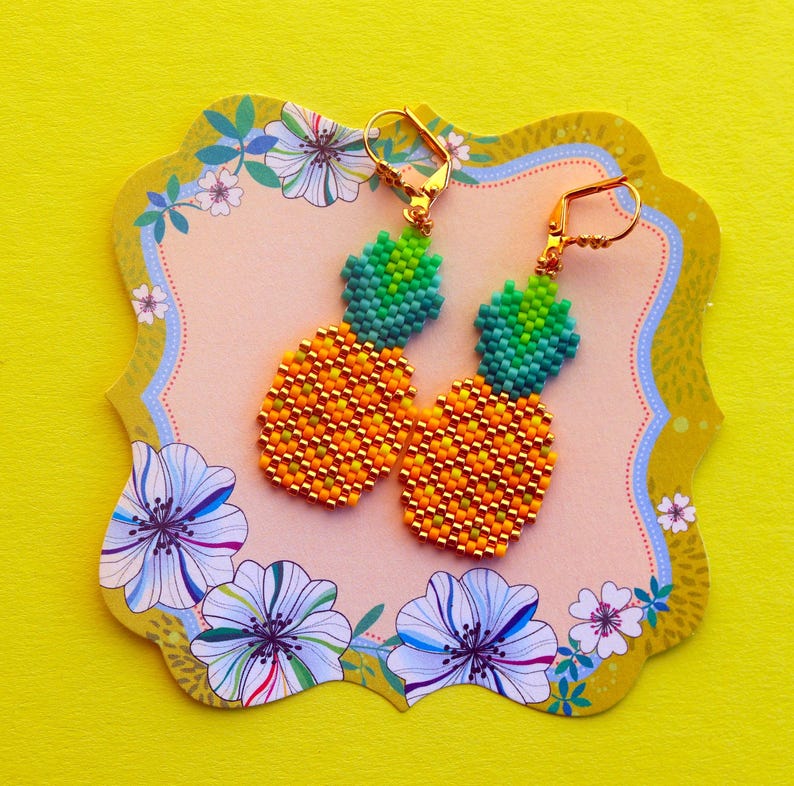 Puede incluir: Un par de pendientes de pi&ntilde;a dorados con hojas verdes, hechos con peque&ntilde;as cuentas. Los pendientes est&aacute;n sobre un fondo rosa con dise&ntilde;os florales.