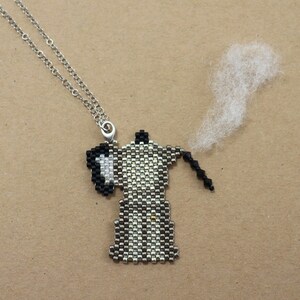 May include: A beaded coffee pot pendant necklace with a silver chain. The pendant is black, white, and silver, with a small wisp of white material coming from the spout. The necklace is on a brown background.