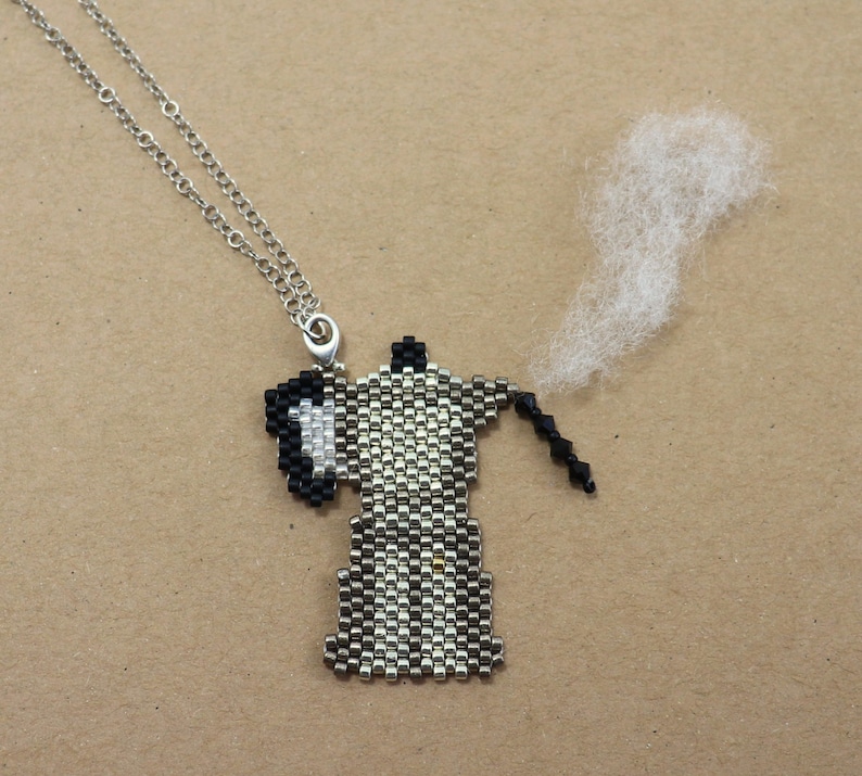 May include: A beaded coffee pot pendant necklace with a silver chain. The pendant is black, white, and silver, with a small wisp of white material coming from the spout. The necklace is on a brown background.