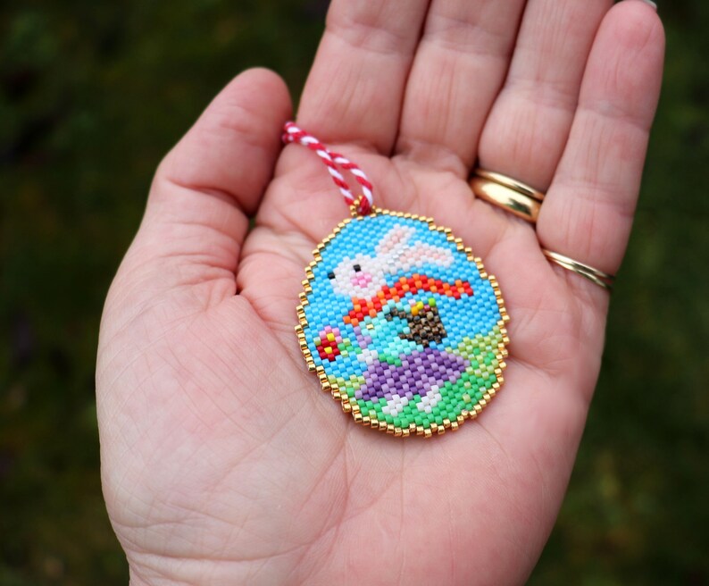 May include: A beaded ornament featuring a white rabbit with a red scarf, surrounded by colourful flowers and greenery on a blue background. The ornament is framed with a gold trim and has a red and white string attached.