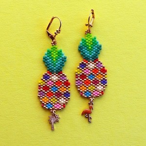 Puede incluir: Un par de pendientes de pi&ntilde;a dorados con cuentas de colores y un peque&ntilde;o encanto de flamenco. Los pendientes est&aacute;n sobre un fondo amarillo.