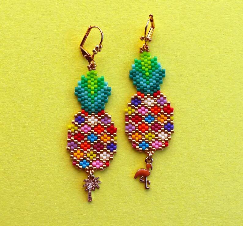 Puede incluir: Un par de pendientes de pi&ntilde;a dorados con cuentas de colores y un peque&ntilde;o encanto de flamenco. Los pendientes est&aacute;n sobre un fondo amarillo.