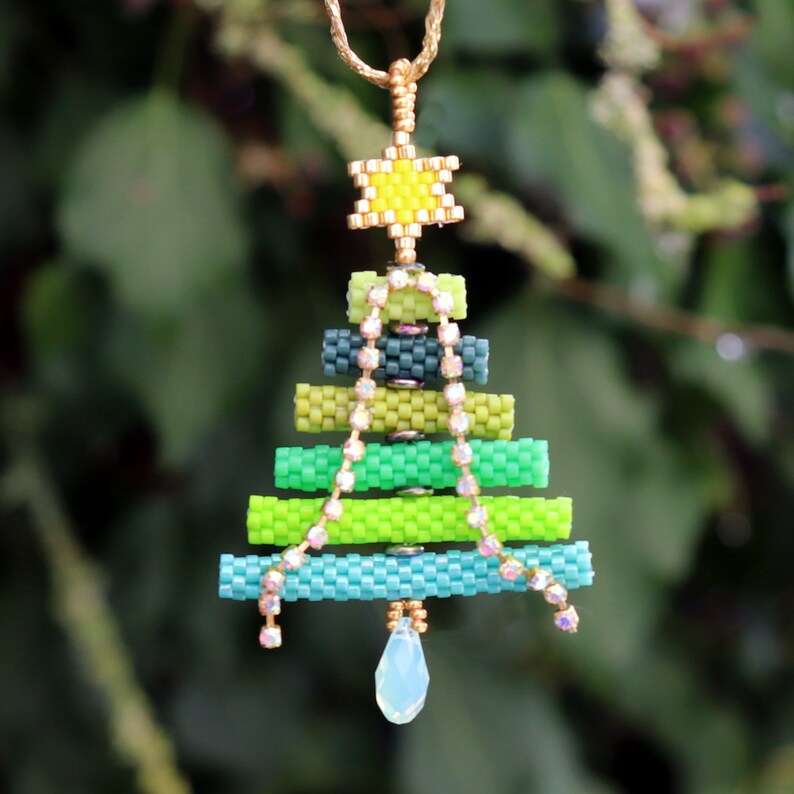 May include: A beaded Christmas tree pendant with a gold chain. The tree is crafted from shades of green and blue beads, topped with a yellow star and a teardrop crystal. A string of crystals hangs down the front, adding sparkle.