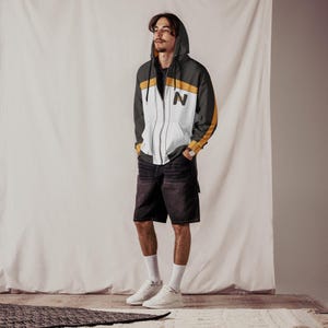 May include: A hooded zip-up jacket with a white front panel, black sleeves, and yellow accents. The jacket features the letter "N" on the chest. The person is wearing black denim shorts, white socks, and white trainers.