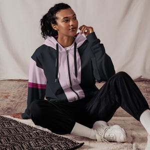 Puede incluir: Una sudadera con cremallera azul marino, rosa claro y magenta. La sudadera con capucha tiene una cremallera de largo completo y una capucha. Las mangas son azul marino con un panel rosa claro y rayas magenta. La persona lleva pantalones negros y zapatillas blancas.