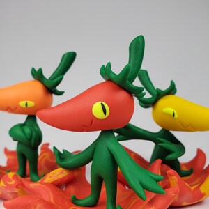 May include: Three whimsical, colorful pepper-shaped figurines with green stems and arms, and yellow eyes. The peppers are orange, red, and yellow, standing on a fiery red and orange base. The background is a plain white.