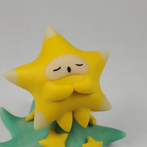 May include: A yellow star-shaped ornament with a peaceful expression, closed eyes, and hands clasped together. The star has white tips and sits on a green base with yellow star details. A decorative item.