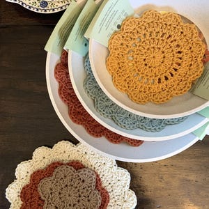 May include: A collection of handmade crocheted coasters in shades of mustard, rust, and sage green, displayed in speckled white ceramic bowls. The bowls have a scalloped edge. A floral patterned plate is in the background. The coasters are for home decor.