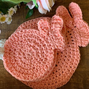 May include: A set of three handmade crochet bunny-shaped items in a soft peach color. The set includes two round coasters and a smaller item with bunny ears. The items are made of a textured yarn and are displayed on a wooden surface.