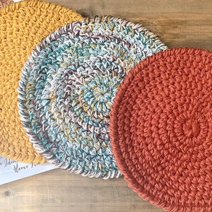 Op de afbeelding: Drie handgemaakte gehaakte onderzetters in tinten geel, blauwgroen en roest. De onderzetters zijn cirkelvormig en hebben een gestructureerd oppervlak. De onderzetters worden waarschijnlijk gebruikt om oppervlakken te beschermen tegen hete gerechten.