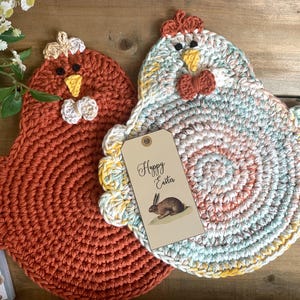May include: Two crocheted chicken pot holders, one in a rust orange color and the other in a multi-colored swirl pattern. The multi-colored pot holder has a tag that says "Happy Easter" with a rabbit illustration.
