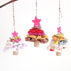 Może przedstawiać: Trzy ręcznie robione ozdoby choinkowe z tkaniny wiszące na gałęzi. Każde drzewko wykonane jest z warstw kolorowej tkaniny i ozdobione różową gwiazdą z filcu. Drzewka są zawieszone na białym sznurku.