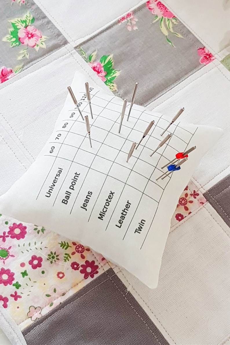 Puede incluir: Un coj&iacute;n de alfileres de tela blanca con una tabla cuadriculada para tama&ntilde;os de agujas de 60 a 110. La tabla est&aacute; etiquetada con diferentes tipos de agujas: Universal, Punta redonda, Jeans, Microtec, Cuero y Gemelas. Hay varias agujas insertadas en el coj&iacute;n de alfileres.