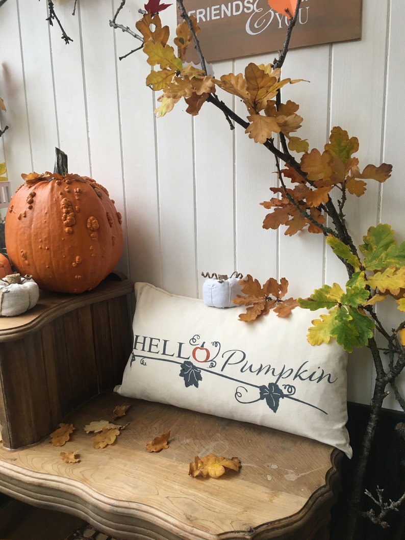Puede incluir: Una almohada blanca con el texto "Hello Pumpkin" en negro y naranja. La almohada est&aacute; sentada en un banco de madera con una calabaza naranja grande y hojas de oto&ntilde;o.