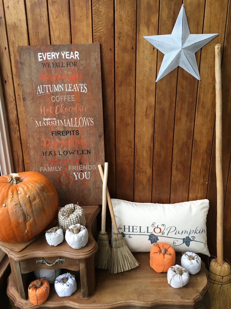 Puede incluir: Un letrero de madera con el texto "Every year we fall for... Autumn leaves, coffee, hot chocolate, marshmallows, firepits, pumpkins, Halloween, family, friends, you". Una calabaza naranja grande, calabazas m&aacute;s peque&ntilde;as y dos escobas est&aacute;n sobre una mesa con una almohada blanca que dice "Hello Pumpkin".