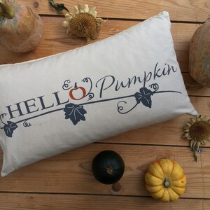 Puede incluir: Una almohada blanca con el texto "HELLO Pumpkin" en letras azules con un peque&ntilde;o dise&ntilde;o de calabaza naranja y vid. La almohada est&aacute; sobre una superficie de madera con peque&ntilde;as calabazas y girasoles.