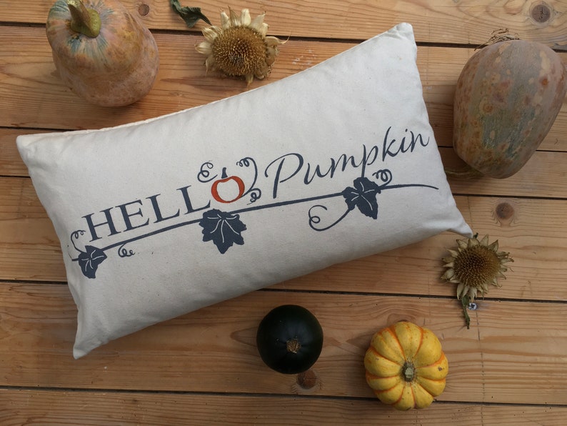Puede incluir: Una almohada blanca con el texto "HELLO Pumpkin" en letras azules con un peque&ntilde;o dise&ntilde;o de calabaza naranja y vid. La almohada est&aacute; sobre una superficie de madera con peque&ntilde;as calabazas y girasoles.