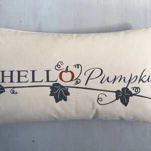 Puede incluir: Una almohada beige con el texto "HELLO Pumpkin" en letras negras con una peque&ntilde;a calabaza naranja y enredaderas de hojas negras.
