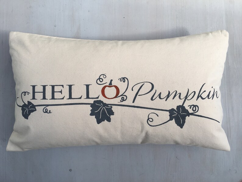 Puede incluir: Una almohada beige con el texto "HELLO Pumpkin" en letras negras con una peque&ntilde;a calabaza naranja y enredaderas de hojas negras.