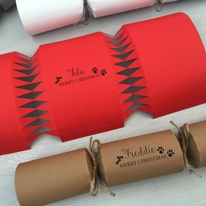 Op de afbeelding: Twee rode en bruine kerstkrakers met de namen "Tala" en "Freddie" erop gedrukt. De crackers hebben een pootprint- en botontwerp en de tekst "Prettige Kerst".