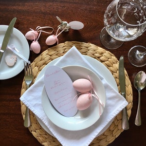 May include: An Easter brunch table setting with white plates, cutlery, and etched glassware. Pink polka-dot eggs with ribbons and a menu card are featured. A woven placemat and wooden table complete the elegant setting.