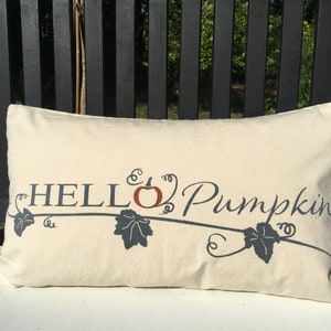Puede incluir: Una almohada blanca con el texto "HELLO Pumpkin" en letras negras con un dise&ntilde;o estilizado de calabaza y vid en naranja y gris.