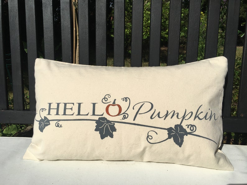 Puede incluir: Una almohada blanca con el texto "HELLO Pumpkin" en letras negras con un dise&ntilde;o estilizado de calabaza y vid en naranja y gris.
