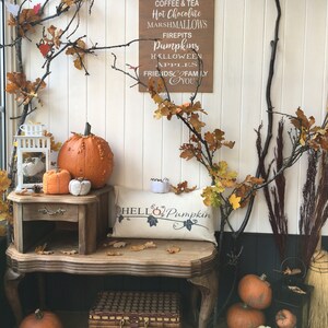 Puede incluir: Un letrero de madera con el texto "Every Year We Fall For Pumpkin Spice, Autumn Leaves, Coffee & Tea, Hot Chocolate, Marshmallows, Firepits, Pumpkins, Halloween Apples, Friends & Family, You". El letrero est&aacute; colgado en una pared blanca con una mesa de madera y calabazas delante. Hay una almohada en la mesa que dice "Hello Pumpkin".
