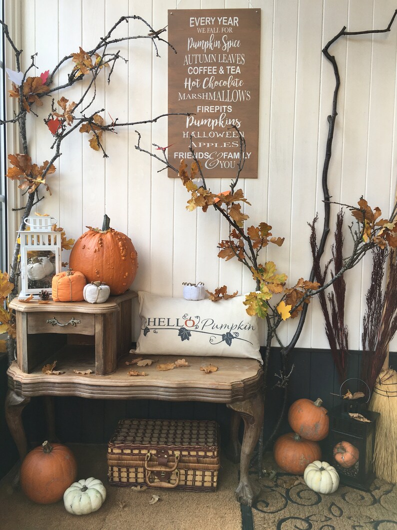 Puede incluir: Un letrero de madera con el texto "Every Year We Fall For Pumpkin Spice, Autumn Leaves, Coffee & Tea, Hot Chocolate, Marshmallows, Firepits, Pumpkins, Halloween Apples, Friends & Family, You". El letrero est&aacute; colgado en una pared blanca con una mesa de madera y calabazas delante. Hay una almohada en la mesa que dice "Hello Pumpkin".