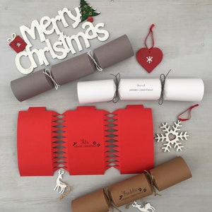 Puede incluir: Una colección de crackers de Navidad personalizados con varios diseños. Los crackers están hechos de papel marrón, papel blanco y papel rojo. Los crackers están decorados con diseños festivos, incluyendo un copo de nieve, un corazón y un reno. Los crackers también están personalizados con nombres, como "Freddie" y "Lola".