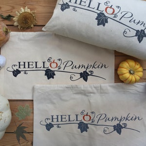 Puede incluir: Tres bolsas de lona blancas con el texto "HELLO Pumpkin" en azul y naranja. Las bolsas est&aacute;n dispuestas sobre una superficie de madera con varias calabazas y follaje de oto&ntilde;o.