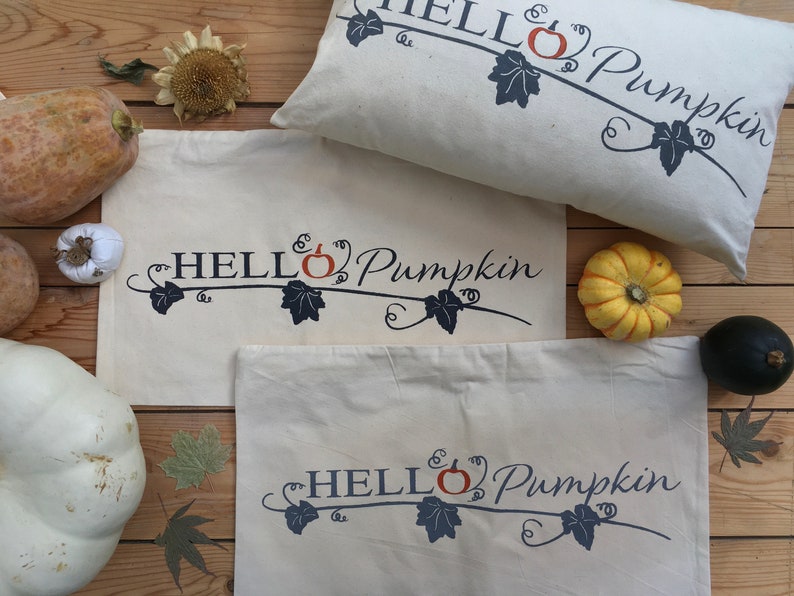 Puede incluir: Tres bolsas de lona blancas con el texto "HELLO Pumpkin" en azul y naranja. Las bolsas est&aacute;n dispuestas sobre una superficie de madera con varias calabazas y follaje de oto&ntilde;o.