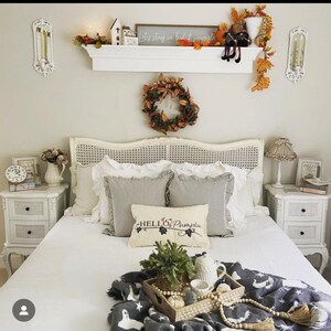 Puede incluir: Una cama blanca con una manta de patr&oacute;n blanco y gris y una almohada con el texto "Hello Pumpkin". La cama est&aacute; decorada con una corona de hojas de oto&ntilde;o, una repisa con velas y un letrero que dice "Let's stay in bed & snuggle", y una bandeja con una planta, una tetera y una taza.