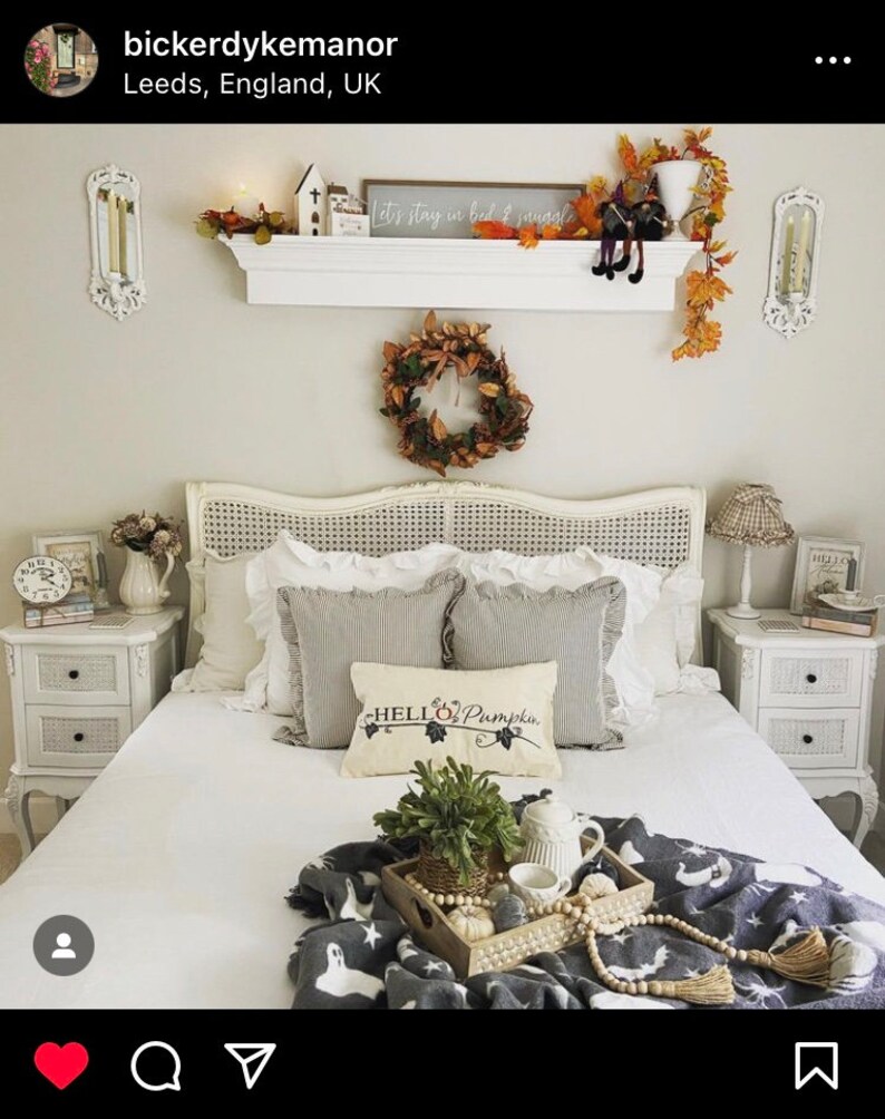 Puede incluir: Una cama blanca con una manta de patr&oacute;n blanco y gris y una almohada con el texto "Hello Pumpkin". La cama est&aacute; decorada con una corona de hojas de oto&ntilde;o, una repisa con velas y un letrero que dice "Let's stay in bed & snuggle", y una bandeja con una planta, una tetera y una taza.