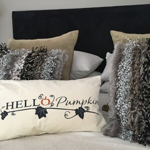 Puede incluir: Una almohada blanca con el texto "HELLO Pumpkin" en negro, con una peque&ntilde;a calabaza naranja y hojas negras. La almohada est&aacute; sobre una cama con una cabecera negra y varias almohadas m&aacute;s en tonos grises y marrones.