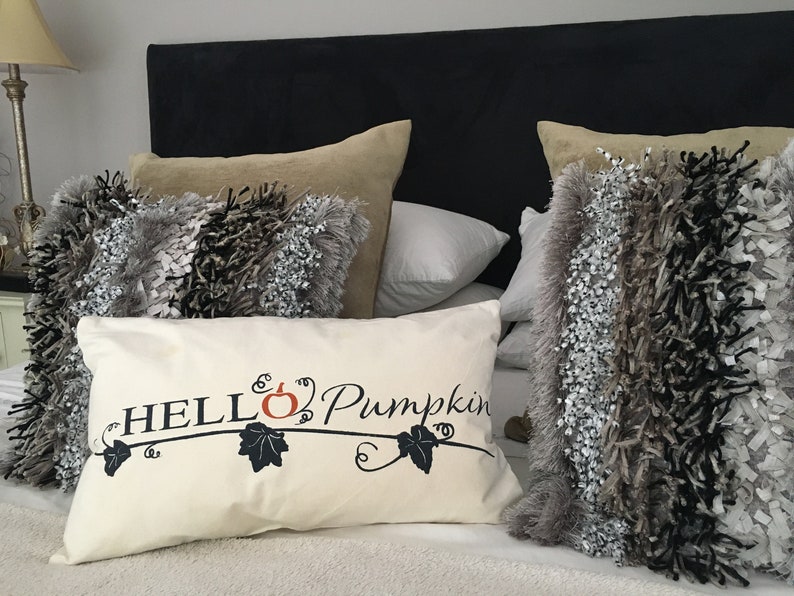 Puede incluir: Una almohada blanca con el texto "HELLO Pumpkin" en negro, con una peque&ntilde;a calabaza naranja y hojas negras. La almohada est&aacute; sobre una cama con una cabecera negra y varias almohadas m&aacute;s en tonos grises y marrones.