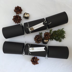 Puede incluir: Dos crackers navideños negros con etiquetas blancas. Las etiquetas tienen el texto "I'll be home for Christmas" y "A Christmas Tree". Los crackers están decorados con pequeñas piñas y campanas plateadas.
