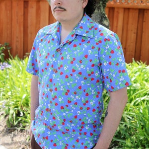 May include: A short-sleeved button-up shirt with a blue background and a pattern of red strawberries, white flowers, and green leaves. The shirt has a classic collar and wooden buttons. The person is wearing brown pants.